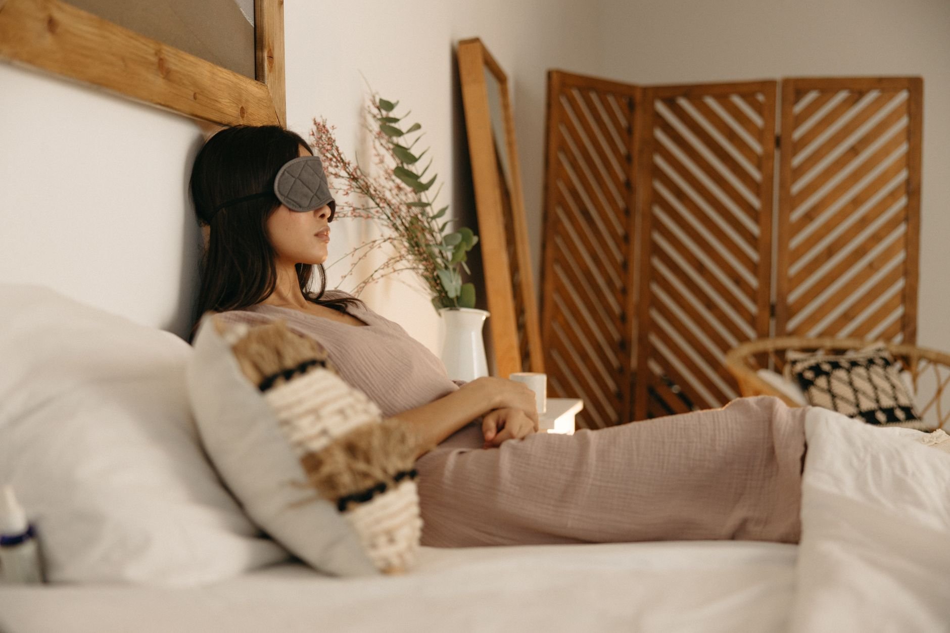 Woman reclining on bed wearing a sleep mask in a cozy, serene bedroom.